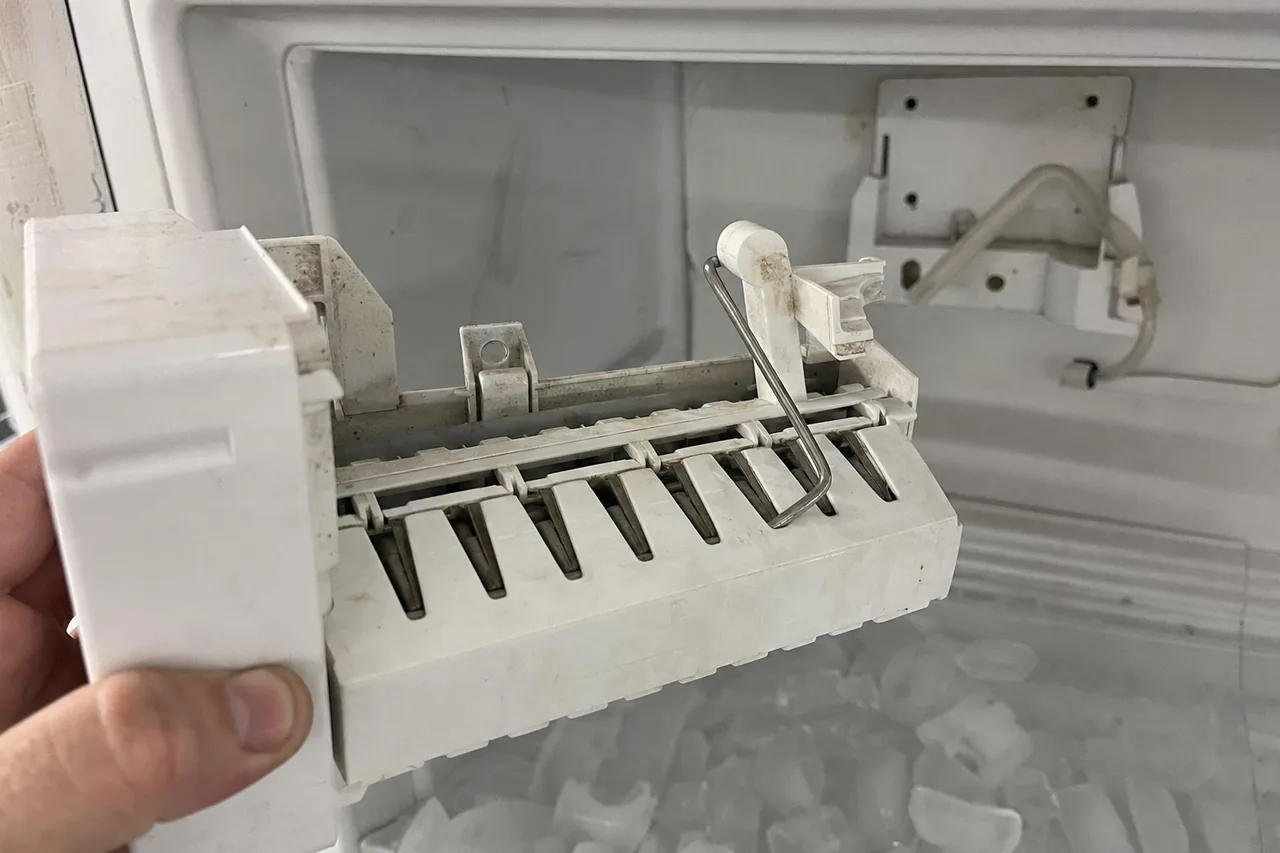 An ice maker module held in one hand clear of the freezer compartment, its harvest arm and white housing visible against the empty mounting bracket.