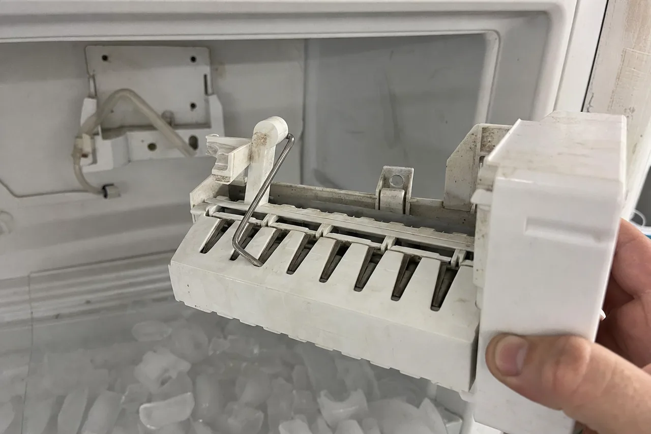 Mirrored view of the ice maker module out of its bracket, water fill tube behind it and melting ice in the bin below.