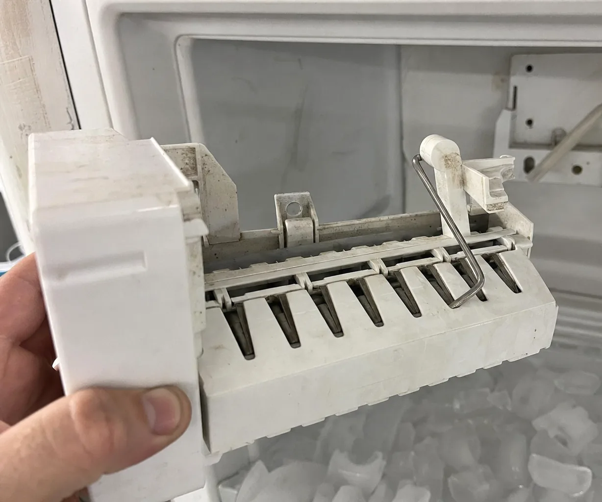 Left crop of the removed ice maker module, harvest arm forward, the freezer wall and its mounting screws behind.