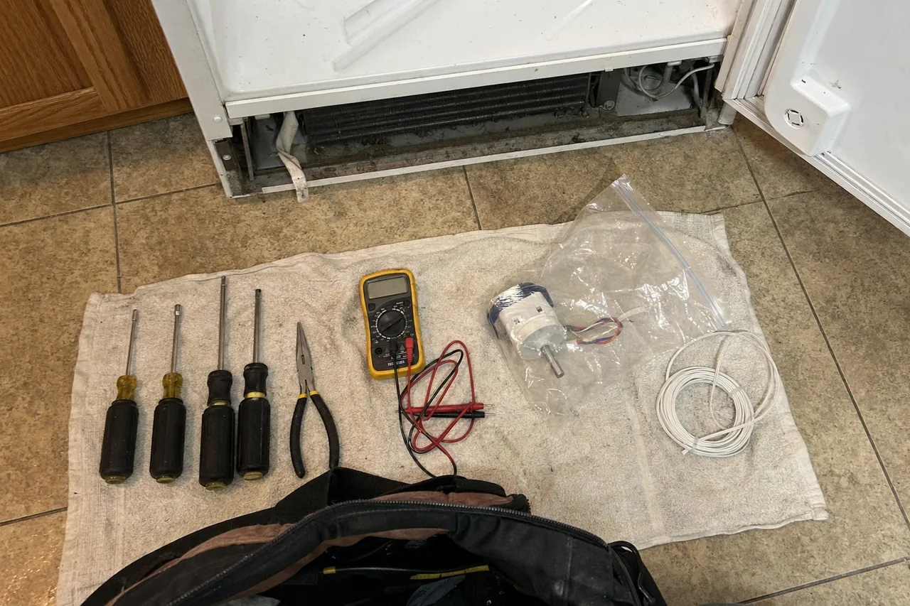 Overhead flat-lay on a folded towel of nut drivers, needle-nose pliers, a digital multimeter and a bagged replacement fan motor, on a kitchen floor by an open refrigerator.